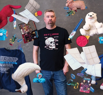 Tim Jones comedic pose with random objects dropping all around him including books, stuffed animals and robotics.